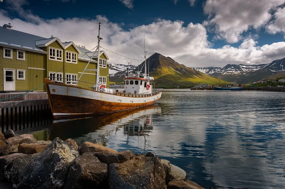 A picturesque Icelandic harbor scene featuring a classic wooden fishing boat docked beside green wooden houses. The water is calm, perfectly reflecting the boat and the surrounding snow-capped mountains in the distance. The sky above is partly cloudy, casting a mix of sunlight and shadows across the landscape, creating a tranquil and scenic atmosphere.