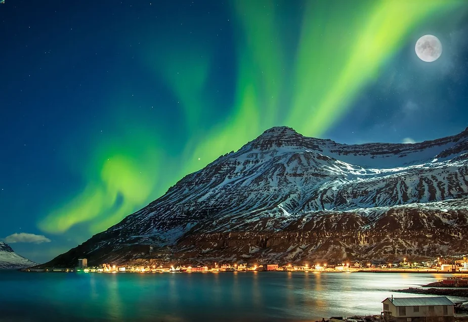 A breathtaking night view of the Northern Lights (Aurora Borealis) dancing across the sky above a snow-capped mountain in Iceland. The vibrant green lights create a mesmerizing contrast against the dark blue sky, with the full moon visible on the right. Below, a small coastal town is softly illuminated, its lights reflecting on the calm waters of a fjord, completing the serene and magical scene.