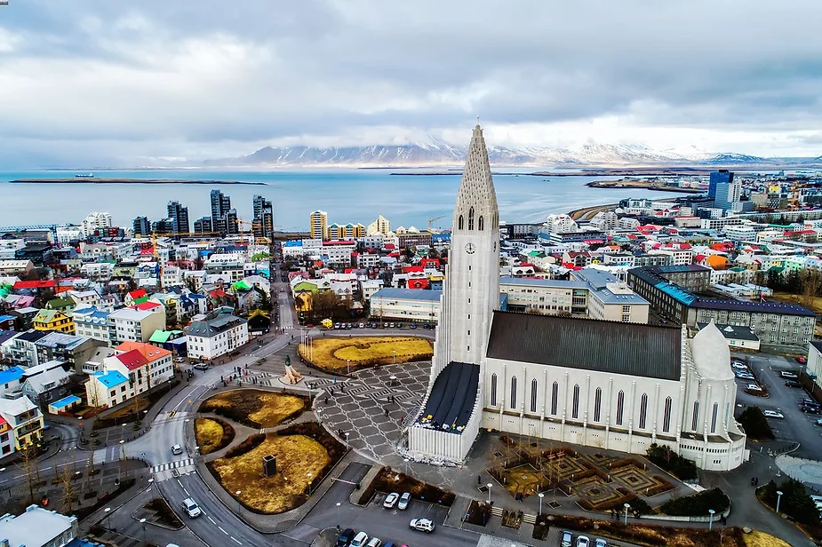 An aerial view of Reykjavik, Iceland, showcasing the iconic Hallgrímskirkja church dominating the skyline with its distinct white tower. The vibrant cityscape is dotted with colorful buildings and modern structures, set against the backdrop of a calm bay and distant snow-covered mountains. The sky is cloudy, but the city's charm shines through in this picturesque landscape.