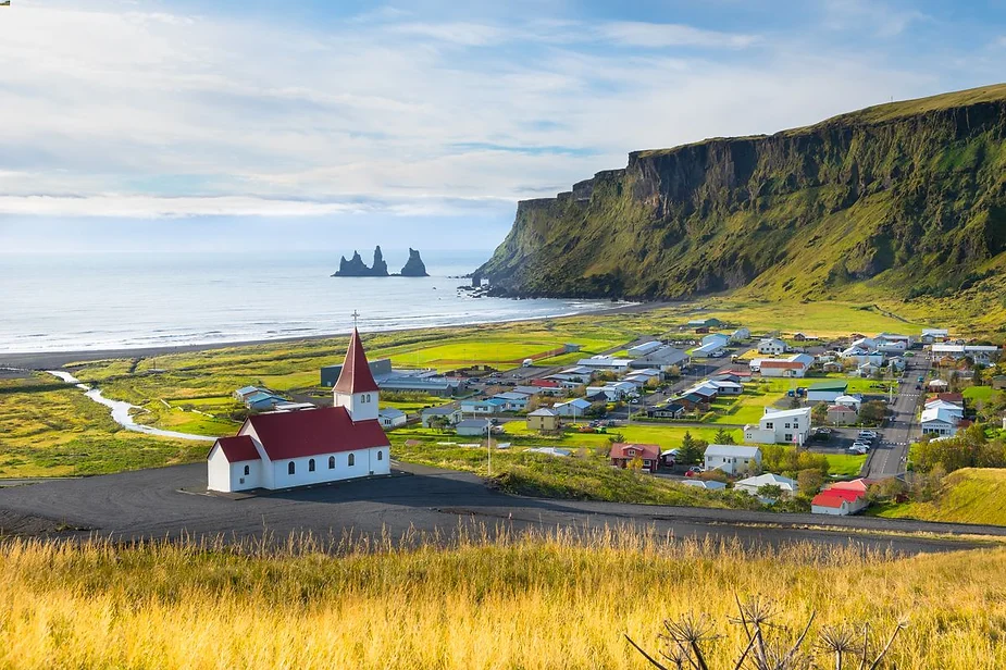 A picturesque view of a small Icelandic village nestled along the coastline, with a charming white church topped with a red roof in the foreground. The village is surrounded by lush green fields, with colorful houses scattered throughout. In the distance, towering cliffs rise dramatically from the shore, and the calm ocean extends to the horizon, where striking rock formations known as Reynisdrangar stand out in the water. The sky is a mix of soft clouds and clear patches, adding to the peaceful atmosphere of the scene.