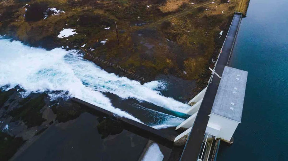 Aerial view of a hydroelectric power station in Iceland. The image shows a powerful stream of water being released from the dam, creating white frothy waves as it flows into the surrounding area. The dam is modern, with a rectangular building structure on one side, and the water is clear and blue, contrasting with the dark, rocky terrain. Patches of snow are visible on the ground, suggesting a cold climate. The surrounding landscape is mostly barren with some vegetation.