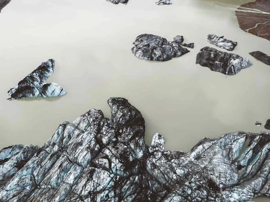 Aerial view of glacial icebergs floating in a milky-colored lake in Iceland. The icebergs are dark with streaks of white and blue, indicating the presence of both ice and volcanic ash. The lake water is a muted, pale brownish-grey, which contrasts with the textured, jagged surfaces of the icebergs. The landscape surrounding the lake is stark and barren, emphasizing the remote and rugged nature of the area.