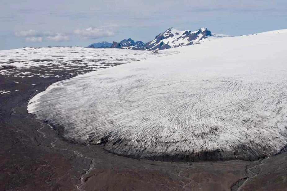 An expansive glacier flows down from a high mountain range in Iceland. The glacier's surface is white with streaks of darker material, likely sediment, and ice. The glacier meets the rocky, barren landscape below, where meltwater streams carve through the dark soil. In the background, rugged peaks partially covered with snow rise against a partly cloudy sky.