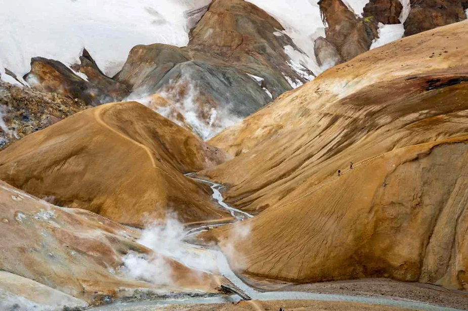 A geothermal landscape in Iceland featuring barren, multicolored hills ranging from ochre to brown, with wisps of steam rising from the earth. The terrain is rugged, with paths winding through the hills, leading the eye to a small stream running through the valley. Snow patches cling to the higher elevations, contrasting sharply with the warm tones of the earth. A couple of tiny figures, likely hikers, can be seen traversing the path, adding a sense of scale to the vast, surreal landscape.