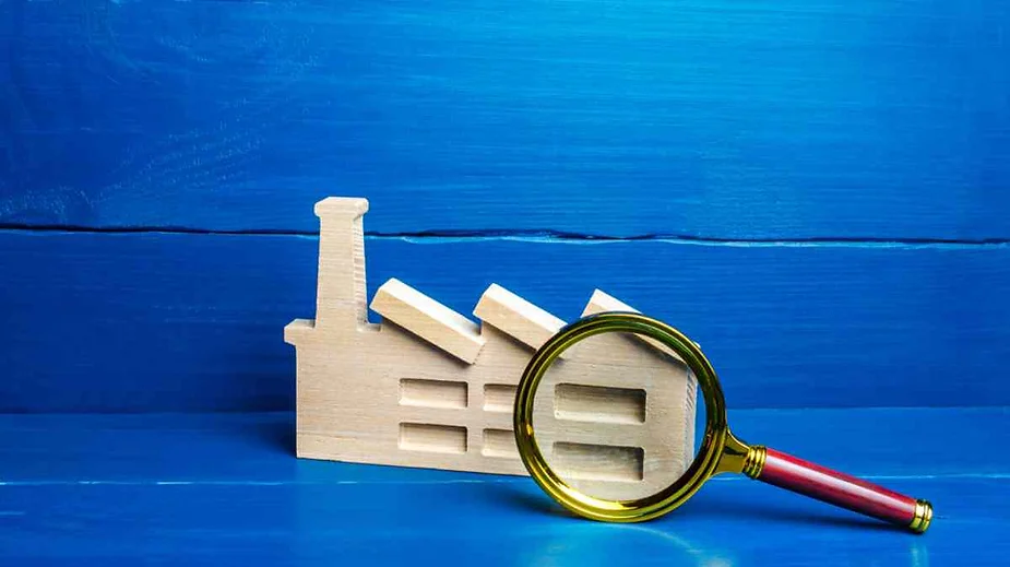 A wooden model of a factory building, characterized by a smokestack and several rectangular windows, is placed on a bright blue wooden surface. Leaning against the model is a magnifying glass with a gold rim and a red handle, suggesting a closer examination or investigation. The background consists of a matching blue wooden wall, creating a uniform and vivid color scheme.