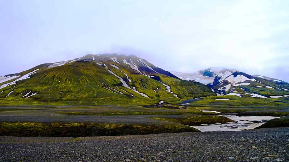 A wide view of rolling hills and mountains in Iceland, partially covered with patches of snow. The hills are lush with green moss and grass, contrasting with the dark volcanic soil below. A small stream cuts through the landscape, meandering around the base of the hills. The sky is overcast, with low clouds partially obscuring the mountain peaks.