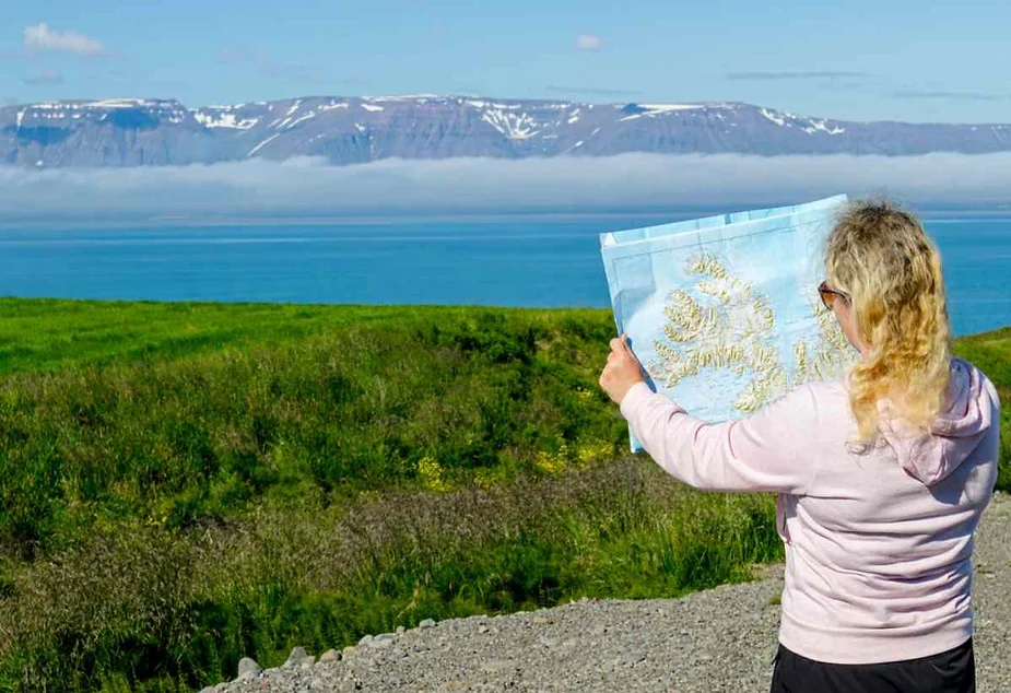 A person with blonde curly hair, wearing a light pink hoodie, is seen from behind as they hold up a map of Iceland. They stand on a gravel path surrounded by lush green grass with wildflowers. In the background, a calm blue sea stretches out, with a layer of low-lying mist or clouds hovering above the water. Beyond the sea, there are snow-capped mountains under a clear blue sky, creating a serene and picturesque scene.