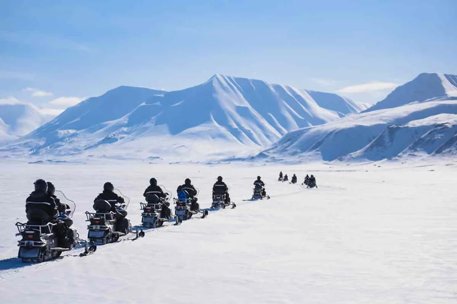 A line of snowmobiles moving across a vast, snowy landscape in Iceland. The snowmobilers, dressed in dark winter gear, form a single file as they head toward the horizon. Towering, snow-covered mountains rise in the background under a clear blue sky, creating a stunning contrast between the riders and the serene, icy expanse. The bright sunlight casts soft shadows on the pristine snow, emphasizing the clarity of the winter day.