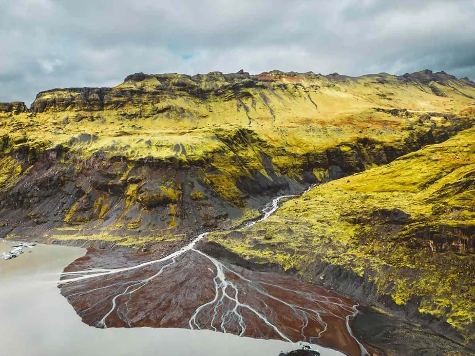 An aerial view of a dramatic landscape in Iceland. The image shows vibrant yellow-green moss covering steep volcanic mountains. Below, a series of braided rivers and streams flow into a muddy, red-tinted delta, which eventually leads into a larger body of water. The contrast between the bright vegetation, dark volcanic rock, and red-brown sediment creates a striking visual effect. The sky is overcast, casting a diffused light over the scene.