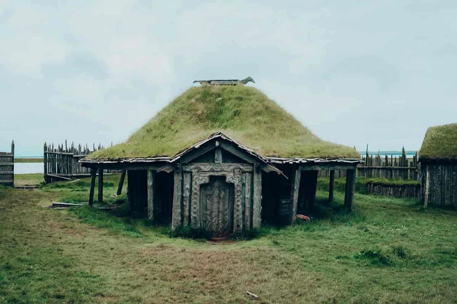 A rustic wooden house with a traditional Viking-style design, featuring a slanted roof covered with grass. The entrance is adorned with intricate carvings, giving it a historic and cultural appearance. The house is situated in a grassy open area, surrounded by a simple wooden fence. The overcast sky adds a moody atmosphere to the scene, emphasizing the connection between the structure and the natural environment.