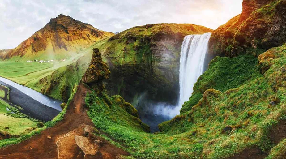 A stunning waterfall cascades down a lush green mountainside in Iceland. The waterfall is surrounded by moss-covered cliffs and vibrant greenery, with a winding river leading away from the falls. The mountains in the background are bathed in a warm, golden light from the setting or rising sun, creating a picturesque and serene landscape. The path in the foreground invites viewers to explore the breathtaking natural beauty of this location.