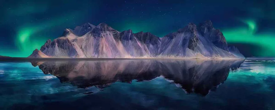 A stunning night scene featuring the Northern Lights (Aurora Borealis) dancing over a range of jagged mountains. The mountains, reflected perfectly in a calm body of water, are bathed in a surreal, ethereal light that ranges from purple to green. The sky is clear, showcasing stars that add to the magical atmosphere, while the vibrant green hues of the Northern Lights contrast beautifully with the deep blues of the night sky and water.