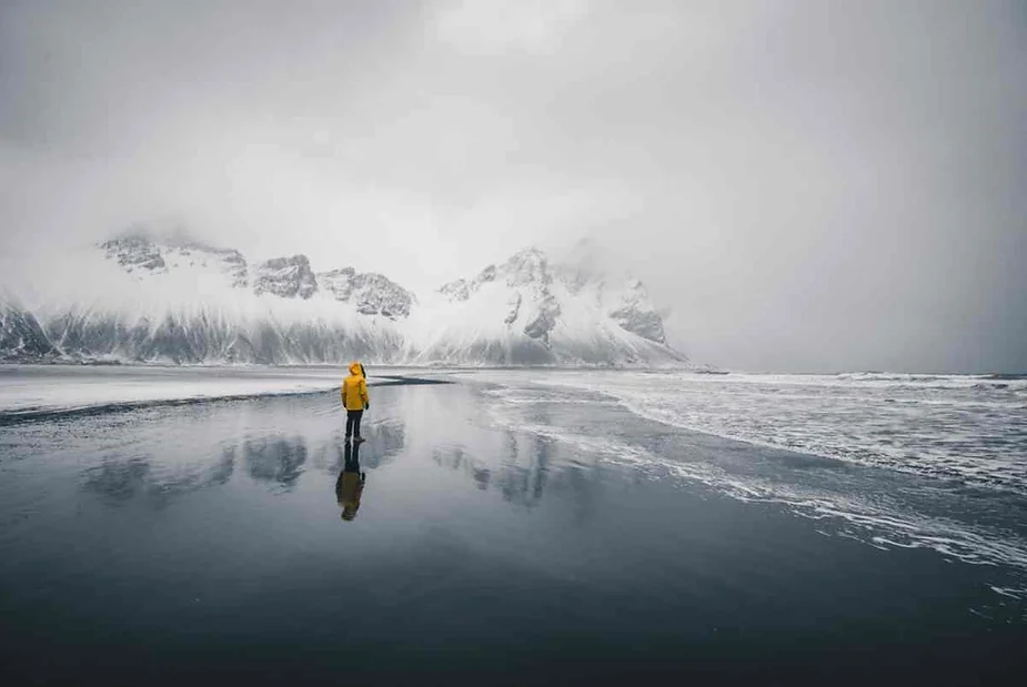 A solitary figure in a bright yellow jacket stands on a vast, reflective black sand beach in Iceland. The scene is dominated by a dramatic mountain range partially covered in snow and shrouded in mist, creating a sense of isolation and grandeur. The cloudy sky adds to the moody atmosphere, while the calm water reflects the mountains and the person, emphasizing the stark contrast between the yellow jacket and the muted, wintry landscape.