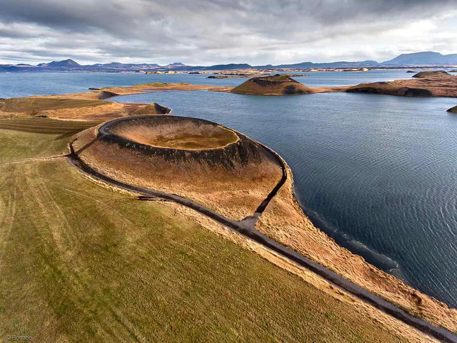 An aerial view of a striking volcanic crater in Iceland, situated near a large body of water. The crater is perfectly circular with a raised rim, and its dark, rocky interior contrasts with the surrounding brown and green landscape. A pathway winds around the crater, leading the eye towards the calm water and other distant volcanic formations. The sky above is cloudy, adding a moody atmosphere to the stark, natural beauty of the scene.