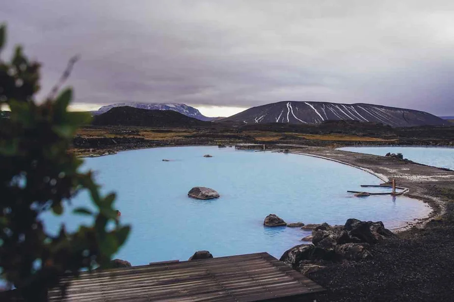 A serene geothermal pool in Iceland with milky blue water, surrounded by dark volcanic rocks and rugged terrain. In the background, a snow-streaked mountain looms under a cloudy, overcast sky. The foreground features a wooden platform leading into the water, with a small bush partially visible on the left side of the image, adding a touch of greenery to the otherwise stark landscape. The scene captures the tranquil beauty of Iceland's natural geothermal features.