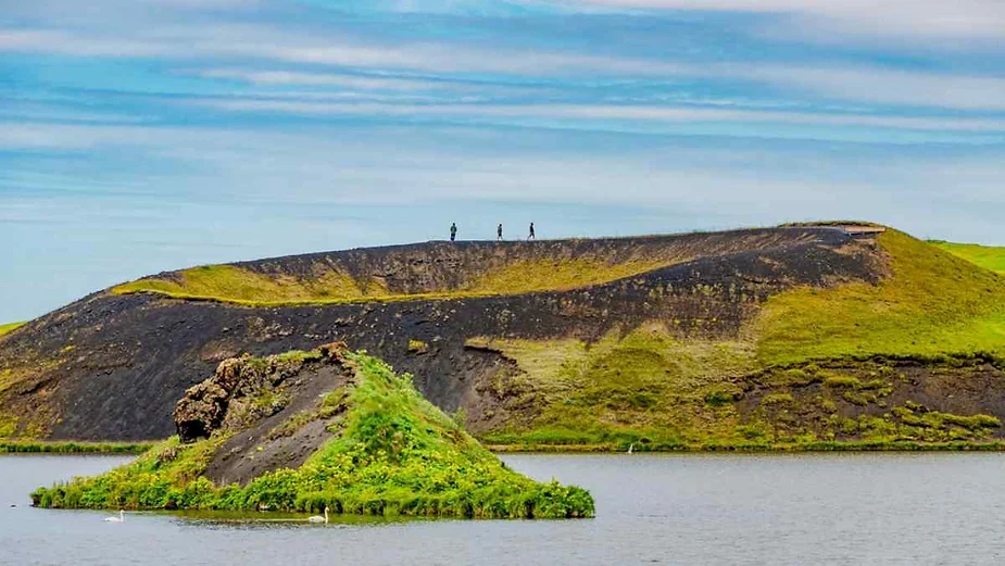 A scenic view of a grassy volcanic crater rising above a lake in Iceland. The crater, which has a dark, rocky surface on one side and is covered with vibrant green grass on the other, creates a striking contrast in the landscape. A small, green, rocky island is visible in the foreground, surrounded by the calm waters of the lake. The sky above is clear with a few soft clouds, and a few tiny human figures can be seen walking along the rim of the crater, giving a sense of scale to the natural feature.