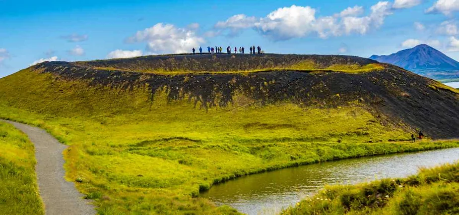 A lush, green volcanic crater in Iceland with a group of people standing on the rim, silhouetted against a bright blue sky dotted with white clouds. The crater's sides are covered with vibrant green grass, and a dark, rocky path winds around the base. In the foreground, a tranquil pond reflects the surrounding landscape, while in the background, another distant mountain rises, adding depth to the scene. The image captures the unique and peaceful beauty of Iceland's volcanic terrain.