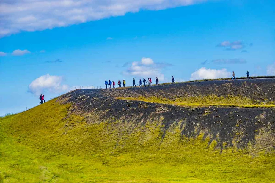 A group of hikers walks along the rim of a volcanic crater in Iceland. The foreground shows the gentle slope of the crater, covered in a mix of green moss and dark volcanic rock. The hikers, dressed in colorful outdoor gear, are silhouetted against a bright blue sky with scattered white clouds. The image captures the scale of the natural landscape and the sense of adventure as the hikers explore this unique geological formation.