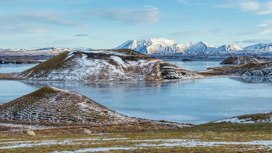 A stunning landscape of icy volcanic craters surrounded by a frozen lake in Iceland. The craters are partially covered in a light dusting of snow, contrasting with the dark, earthy tones of the land. In the background, majestic snow-capped mountains rise under a clear blue sky, adding depth to the scene. The serene frozen lake mirrors the cold yet beautiful Icelandic winter environment, with patches of brown grass visible in the foreground.