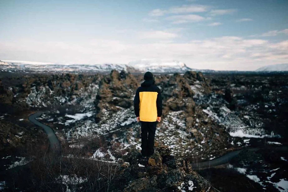 A person wearing a yellow and black jacket stands on the edge of a rocky outcrop, looking out over a rugged, snow-dusted volcanic landscape in Iceland. The winding road below curves through the terrain, leading the eye towards distant snow-capped mountains under a partly cloudy sky. The image captures the vastness of the landscape and the solitude of the figure, emphasizing the dramatic and stark beauty of the Icelandic wilderness.