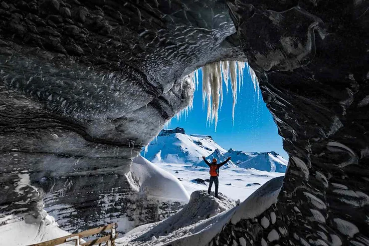 A hiker stands triumphantly at the entrance of the Katla Ice Cave in Iceland, with arms raised against a stunning backdrop of snow-covered mountains and a bright blue sky. The cave's interior is framed by rugged, icy formations, and sharp icicles hang from the roof, capturing the raw beauty of the frozen landscape. The hiker, dressed in a bright orange jacket, adds a vibrant contrast to the predominantly blue and white surroundings. This image encapsulates the sense of adventure and awe inspired by exploring Iceland's majestic ice caves.