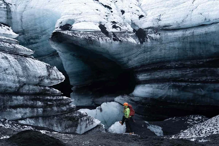 A hiker stands in front of the entrance to the Katla Ice Cave in Iceland. The cave is surrounded by layers of dark volcanic ash and thick, blue-tinted ice, creating a dramatic contrast. The hiker, wearing a bright green jacket and an orange helmet, appears small against the massive ice formation, highlighting the cave's impressive scale. The entrance to the cave is wide and dark, hinting at the vast, icy world inside. The scene is a testament to the rugged and awe-inspiring natural beauty of Iceland's glacial landscapes.