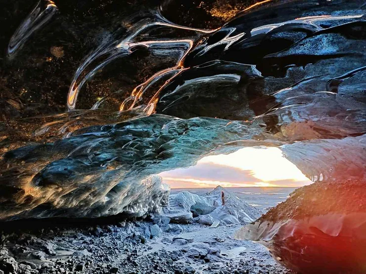 View from inside the Katla Ice Cave in Iceland, looking out towards the horizon. The cave's ceiling is made of smooth, translucent ice, with intricate patterns and textures reflecting light from the outside. The ice has a blue-green tint, creating a striking contrast with the warm, golden light of the sunset visible at the cave's entrance. The ground is covered with rocks and ice, adding to the rugged, natural beauty of the scene. The image captures the serene yet powerful atmosphere of the ice cave and its connection to the vast, frozen landscape beyond.