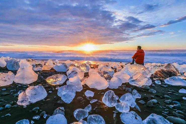 A serene sunrise view at Diamond Beach in Iceland, where large chunks of clear ice, shaped like diamonds, are scattered across the black volcanic sand. The ice pieces glisten under the rising sun, which casts a golden light over the entire scene. A solitary figure, clad in a bright red jacket, sits on one of the larger ice blocks, gazing out at the distant horizon where the sun meets the calm, reflective ocean. The sky is painted with soft hues of orange, pink, and blue, adding to the tranquil and magical atmosphere of this unique location.