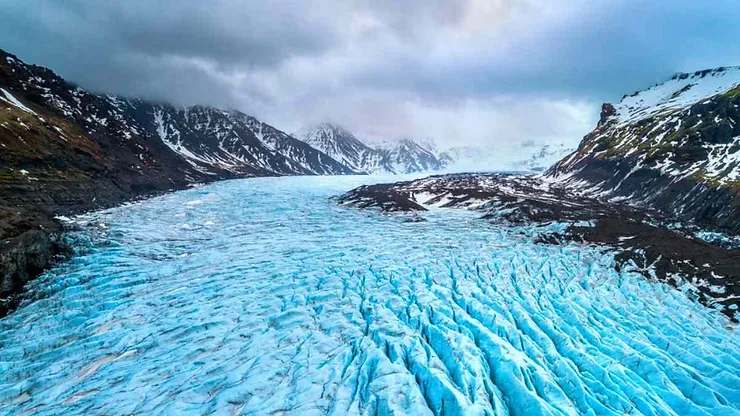 A stunning aerial view of the Vatnajökull Glacier in Iceland, part of the Vatnajökull National Park. The glacier's surface is covered in bright blue ice crevices, stretching out between two rugged mountain ranges. The mountains, capped with snow, rise sharply on either side of the glacier, with some peaks disappearing into the thick, low-hanging clouds. The dark, rocky terrain contrasts with the vivid blue ice, creating a dramatic and awe-inspiring landscape. The overall atmosphere is one of icy grandeur, capturing the raw and powerful beauty of Iceland's largest glacier.