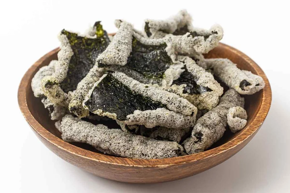 A wooden bowl filled with crispy, puffed snacks that appear to be dried fish skins. The fish skins are grayish in color with a slightly greenish tint from possibly being seasoned with seaweed. The texture looks light and crunchy, with some pieces curled up and others lying flat in the bowl. The background is plain white, putting the focus entirely on the snack.