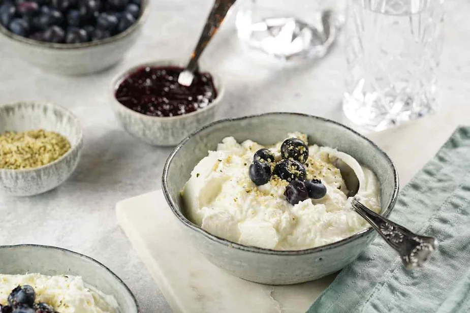 A bowl of creamy yogurt topped with fresh blueberries and sprinkled with a small amount of crushed nuts or seeds. The bowl is accompanied by a vintage-style spoon resting on its side. Surrounding the main dish are additional small bowls containing ingredients like blueberry jam, fresh blueberries, and more crushed nuts or seeds, all placed on a soft white surface. The scene is set with a light, airy atmosphere, complemented by a folded pastel green napkin and a glass of water, suggesting a wholesome and simple breakfast or snack.