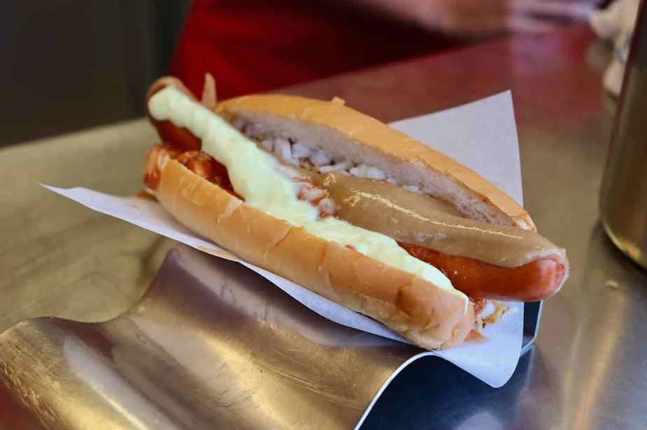 A hot dog served in a soft bun with various toppings. The hot dog is topped with chopped onions, a drizzle of white sauce, and a layer of mustard or a similar sauce. The hot dog is placed on a piece of paper, resting on a shiny metal surface, with a blurred background suggesting a busy food stand or kitchen setting.