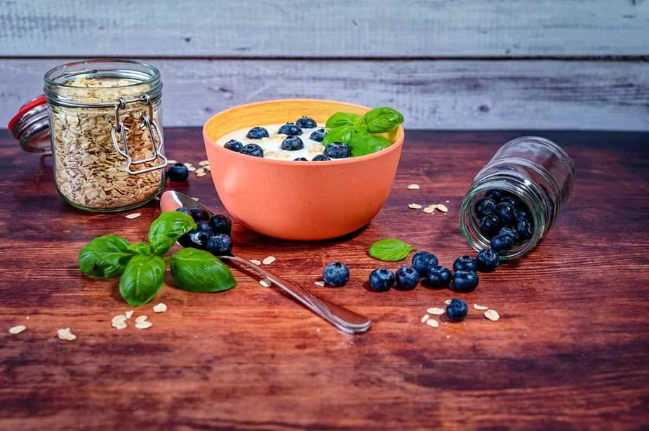 A breakfast scene featuring a bowl of oatmeal topped with fresh blueberries and basil leaves, placed on a rustic wooden table. To the left of the bowl, there's a glass jar filled with rolled oats, partially open. To the right, a jar of blueberries is tipped over, spilling a few berries onto the table. A sprig of fresh basil and a spoon lie beside the bowl, completing the wholesome and fresh presentation.