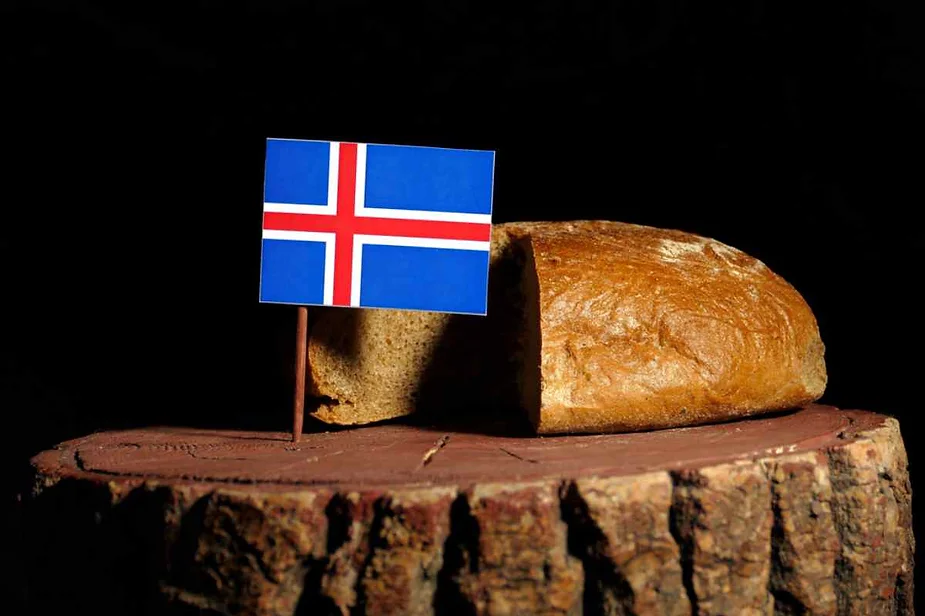 A loaf of rustic bread sits on a wooden surface, likely a tree stump or slab, with a dark background that contrasts the warm tones of the bread. Sticking out from the loaf is a small Icelandic flag on a toothpick, symbolizing the bread's Icelandic origin or significance. The lighting emphasizes the texture of the bread's crust and the wood grain of the stump, creating a simple yet striking image that highlights traditional Icelandic food.