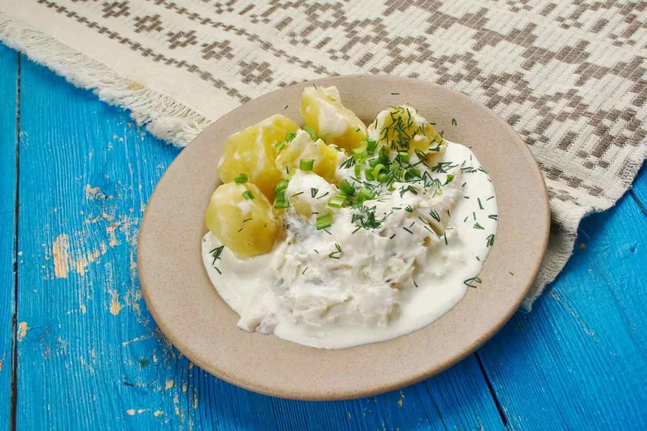 A plate of traditional Icelandic dish is served on a simple beige plate, set on a vibrant blue wooden table. The dish consists of boiled potatoes and fish, smothered in a creamy white sauce, possibly featuring a yogurt or dairy base. The meal is garnished with fresh green herbs like dill and green onions, adding a touch of color to the dish. To the side, there is a woven cloth with a geometric pattern, enhancing the rustic and homey presentation of the meal.