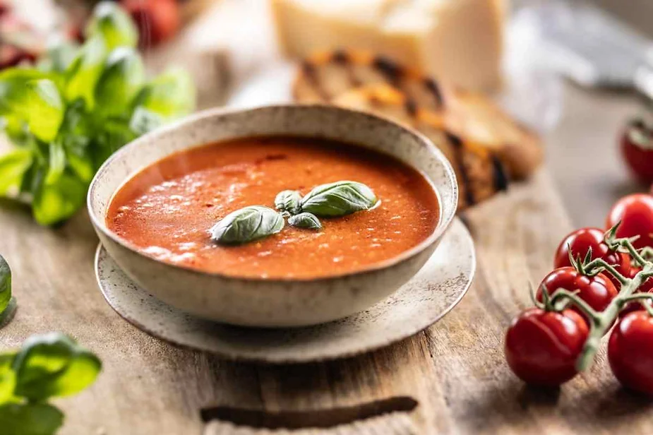 A ceramic bowl filled with a rich, red tomato soup, garnished with a few fresh basil leaves floating on top. The bowl is placed on a matching saucer on a rustic wooden table. Surrounding the bowl are vibrant green basil leaves, a bunch of ripe cherry tomatoes on the vine, and slices of toasted bread in the background, suggesting a cozy meal setting.