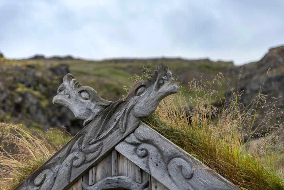 A close-up view of the decorative wooden carvings on the roof of a rustic hut. The carvings depict two stylized dragon or serpent heads emerging from the roof's peak. The wooden structure is weathered, with tufts of grass growing on the thatched roof. The background shows a blurry, grassy hillside under a cloudy sky.