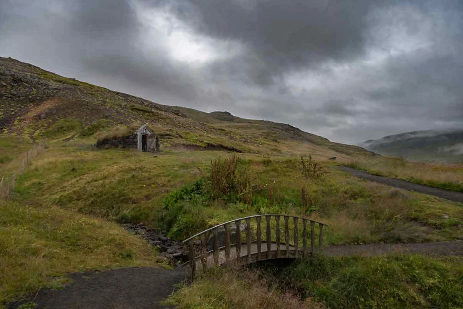 A moody, overcast landscape featuring a small, wooden bridge over a narrow stream. The bridge leads to a dirt path that winds through a grassy, hilly terrain. In the distance, a rustic stone and wooden hut sits nestled against the hillside. The sky is filled with dark, heavy clouds, adding a dramatic atmosphere to the scene.