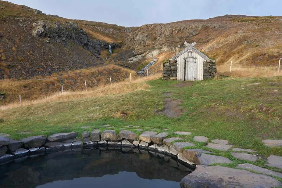 A serene scene featuring a small, stone-lined hot spring pool in the foreground, surrounded by a stone path. In the background, there is a rustic stone and wood hut with a thatched roof, nestled into a grassy hillside. A small waterfall can be seen flowing down the hill behind the hut, adding to the peaceful atmosphere.