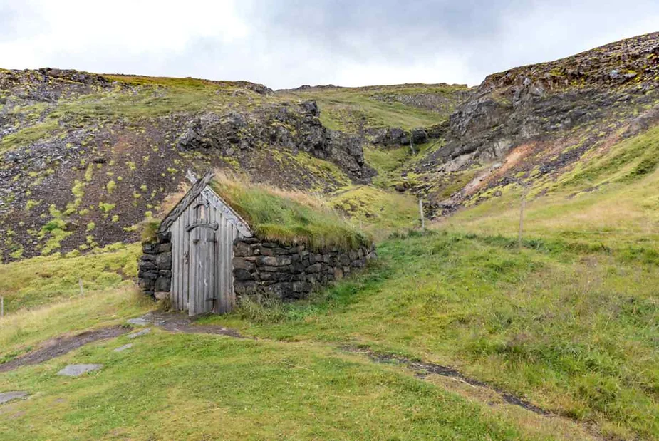 A small, rustic stone and wood hut with a grass-covered roof, situated on a grassy hillside. The landscape surrounding the hut is rugged, with rocky outcrops and a steep hill in the background. The grass is lush and green, contrasting with the darker stone of the hut and the hill.