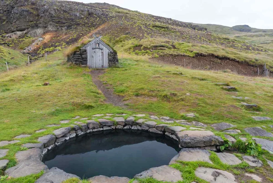 A peaceful outdoor setting featuring a small, circular hot spring pool surrounded by stones. The pool is nestled in a grassy area with a quaint, rustic stone and wooden hut in the background. The hut is connected to the pool by a dirt path. The surrounding landscape is lush with green hills and rugged terrain, creating a serene and natural atmosphere.