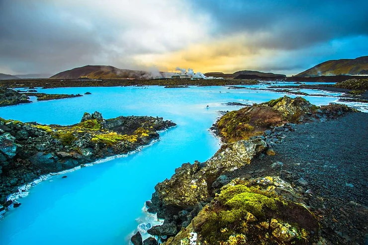 The Blue Lagoon in Iceland, featuring vibrant blue geothermal waters surrounded by rugged volcanic rocks and distant mountains under a dramatic sky.