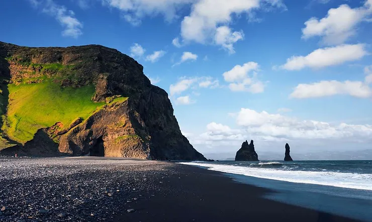 A breathtaking view of Iceland's Black Sand Beach with its iconic volcanic black sand contrasting against the bright blue sky. The shoreline curves along the base of a rugged, moss-covered cliff, with a cave opening visible at its base. Out in the ocean, the Reynisdrangar basalt sea stacks rise dramatically from the water, adding to the mystical atmosphere of this unique landscape.