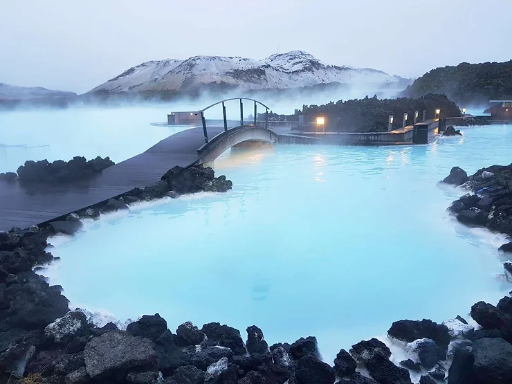 A serene view of the Blue Lagoon in Iceland, with milky blue geothermal waters surrounded by dark volcanic rocks. A small footbridge arches over the lagoon, and steam rises from the warm waters, creating a mystical atmosphere. Snow-covered hills in the background add to the peaceful, wintery scene.