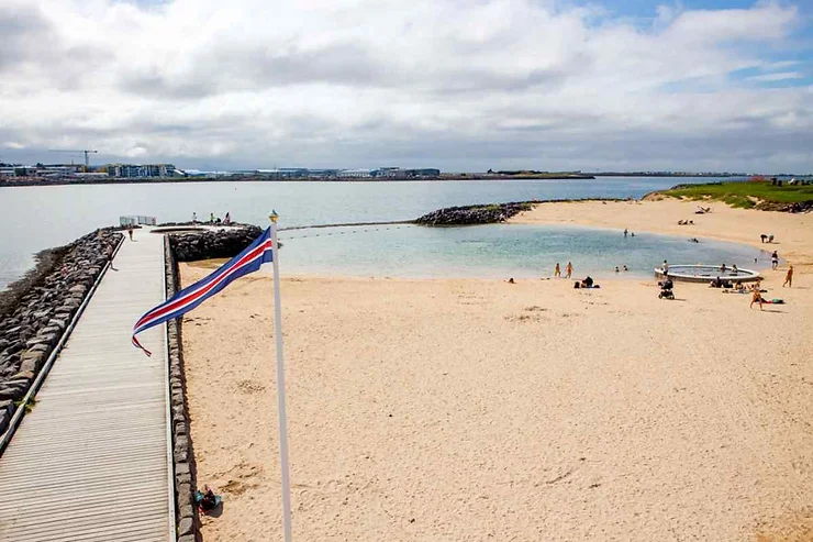 Nauthólsvík Geothermal Beach in Reykjavik, Iceland, showcasing a serene sandy shore, calm waters, and an Icelandic flag waving on a boardwalk.