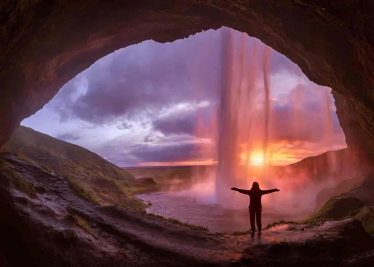 Person standing behind Seljalandsfoss Waterfall in Iceland at sunset, with arms outstretched, capturing the vibrant colors and dramatic sky