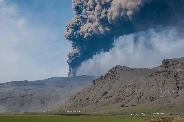 Eyjafjallajökull volcano in Iceland erupting, with a massive ash cloud rising over rugged mountains and small farms in the foreground.