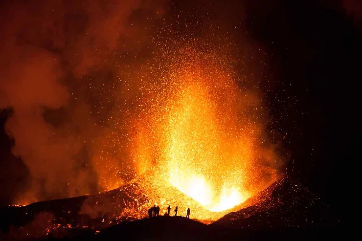 A dramatic night-time image of an active volcanic eruption at Eyjafjallajökull in Iceland. The volcano spews out bright orange lava, with molten rock and ash being violently ejected into the dark sky. Silhouetted against the intense glow of the eruption, a few small figures, possibly onlookers or scientists, stand at a distance on a ridge, witnessing the raw and fiery spectacle. The contrast between the dark surroundings and the fiery eruption emphasizes the incredible power and danger of the natural event, capturing a moment of awe and wonder as nature unleashes its force.