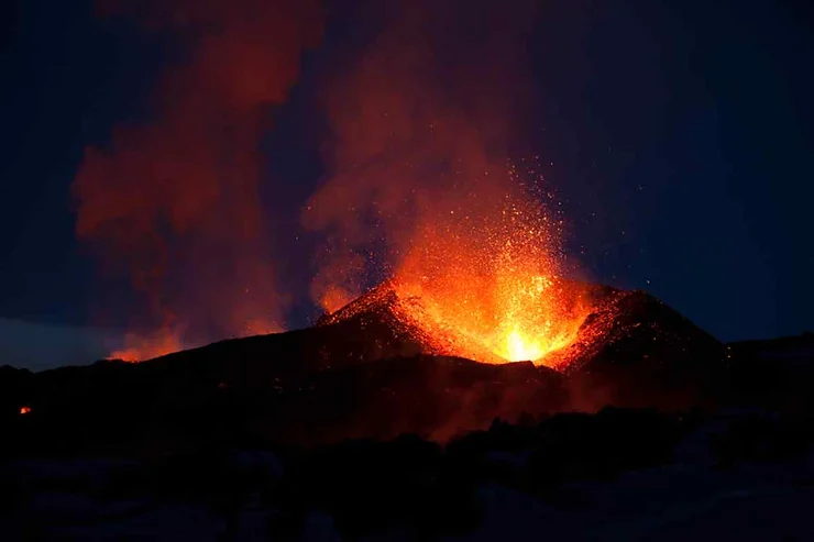 A volcanic eruption in Iceland during the night, with fiery lava spewing from the crater into the dark sky. The eruption casts an intense orange-red glow, illuminating the surrounding smoke and ash plumes that rise into the night. The silhouette of the volcanic cone is visible against the glowing sky, highlighting the dramatic and powerful natural phenomenon. The image captures the raw energy and explosive force of a volcanic eruption, emphasizing the contrast between the bright, molten lava and the dark, shadowed landscape.