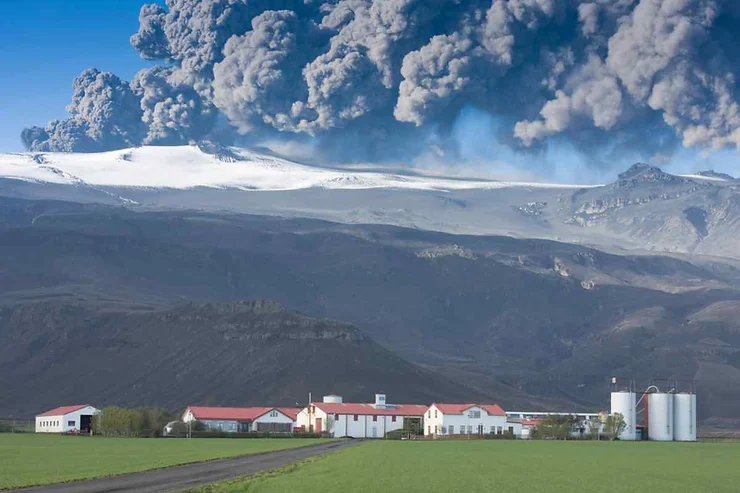 A powerful volcanic eruption at Eyjafjallajökull in Iceland, with massive plumes of ash and smoke billowing high into the sky above the snow-covered volcano. In the foreground, a serene farm with red-roofed buildings and green fields contrasts sharply with the dramatic eruption in the background. The image captures the juxtaposition of calm rural life against the intense and overwhelming force of nature, showcasing the raw power of the volcano as it disrupts the tranquil Icelandic landscape.